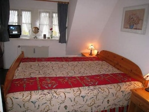 Ein Schlafzimmer mit Bett, Nachttisch, Lampe, einem Fenster mit Vorhängen, einer Heizung und einem Fernseher.
