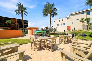 An outdoor patio area with wooden tables and chairs, surrounded by palm trees and a building with multiple windows. - Gulf + Bay Views · Boat Dock · Bradenton Beach (Bradenton Beach)