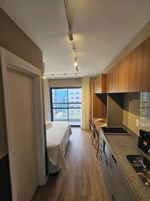 Habitación de hotel compacta con kitchenette, una cama y vista a la ciudad a través de grandes ventanales.