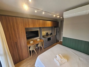 A hotel room with a bed, a desk with a TV, a kitchenette, and an air conditioning unit. - Apartamento Conforto e Charme na Vila Cordeiro (São Paulo)