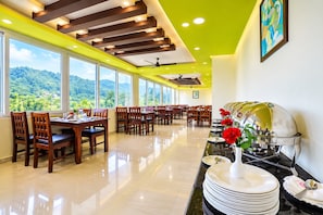 A hotel dining area with multiple tables and chairs, a buffet station with plates and cutlery, and large windows offering a view of the outdoors. - Anglo French Resort Yercaud (Yercaud)