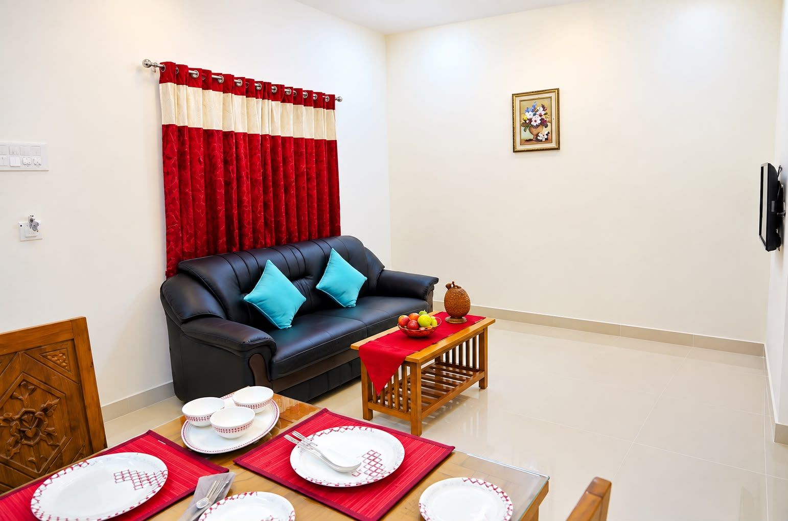 A living room with a black leather sofa, red and white curtains, a small table with fruit, and a wall-mounted TV.