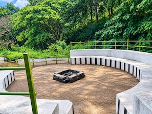 Una estructura circular de concreto con un brasero en el centro, rodeada por una valla metálica verde y vegetación densa.