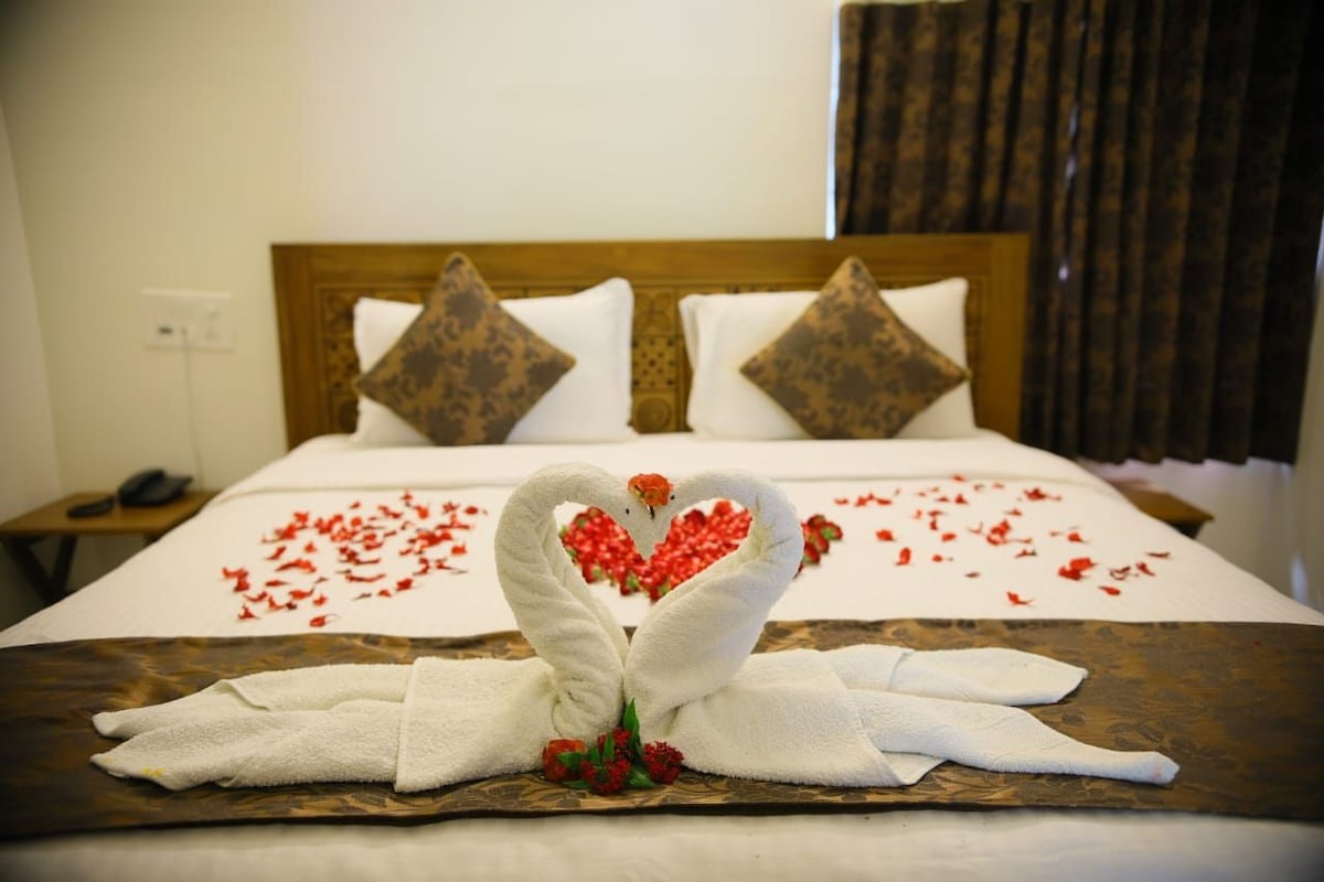 A bed with a heart-shaped towel arrangement and red petals, a wooden headboard, and a nightstand with a phone.