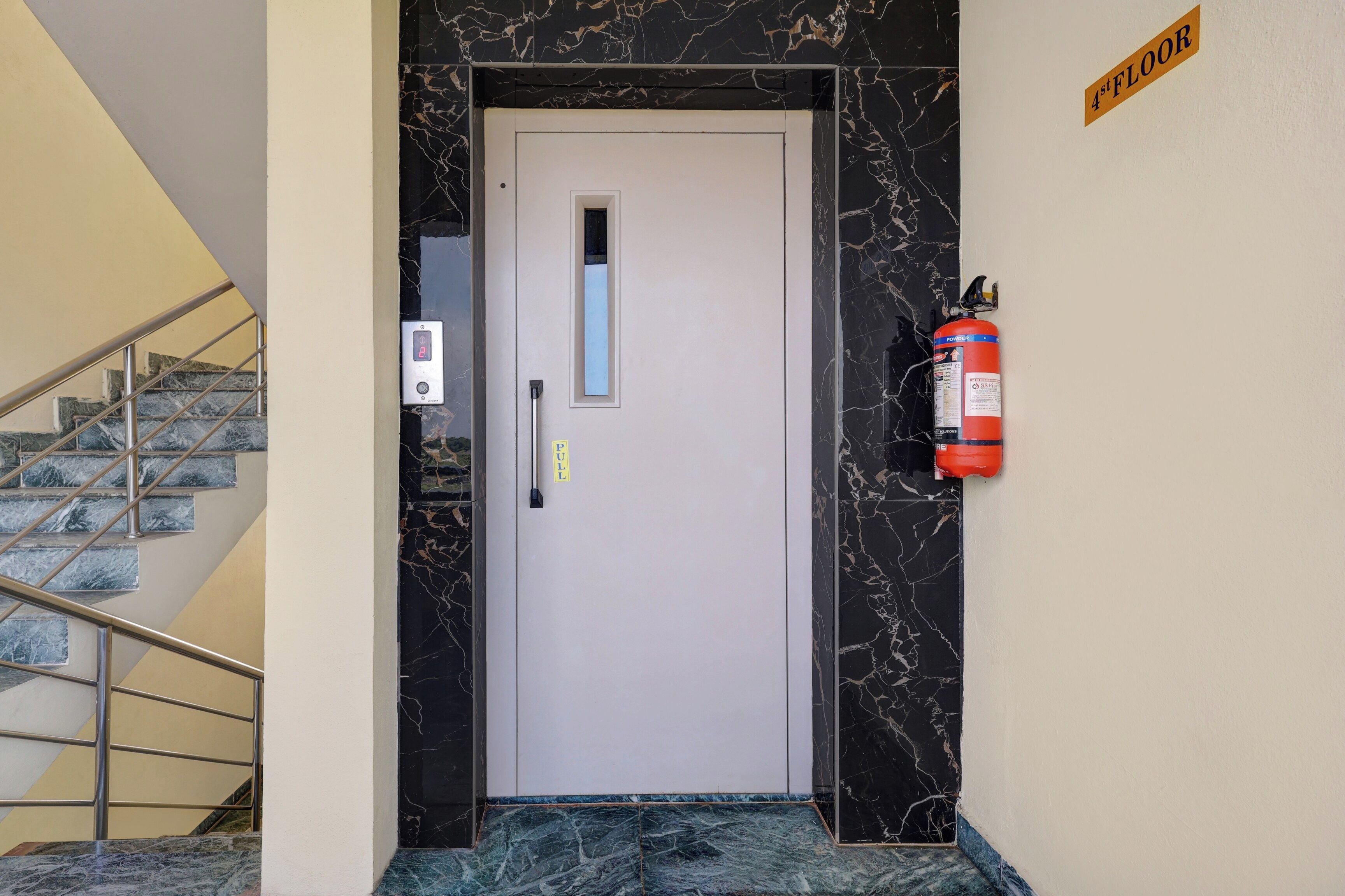 Ein Hotel Eingang mit einer Marmortreppe, einem digitalen Aufzugbedienteil und einem an der Wand befestigten Feuerlöscher.