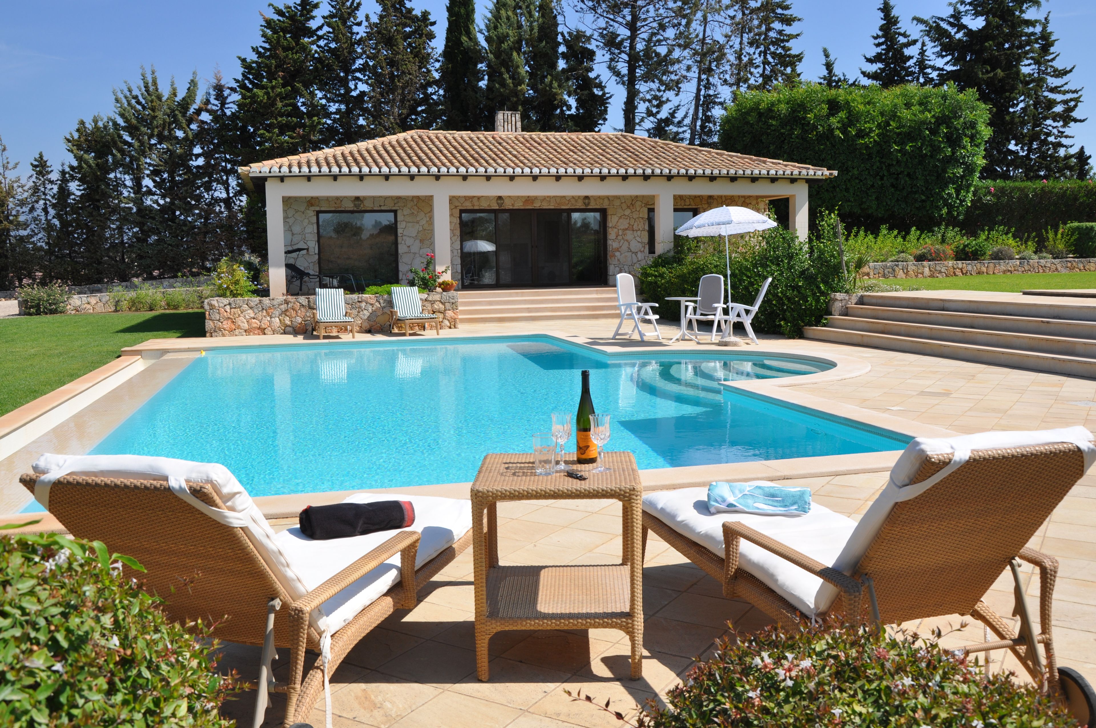 A swimming pool with lounge chairs and a table, surrounded by a stone wall and a garden.