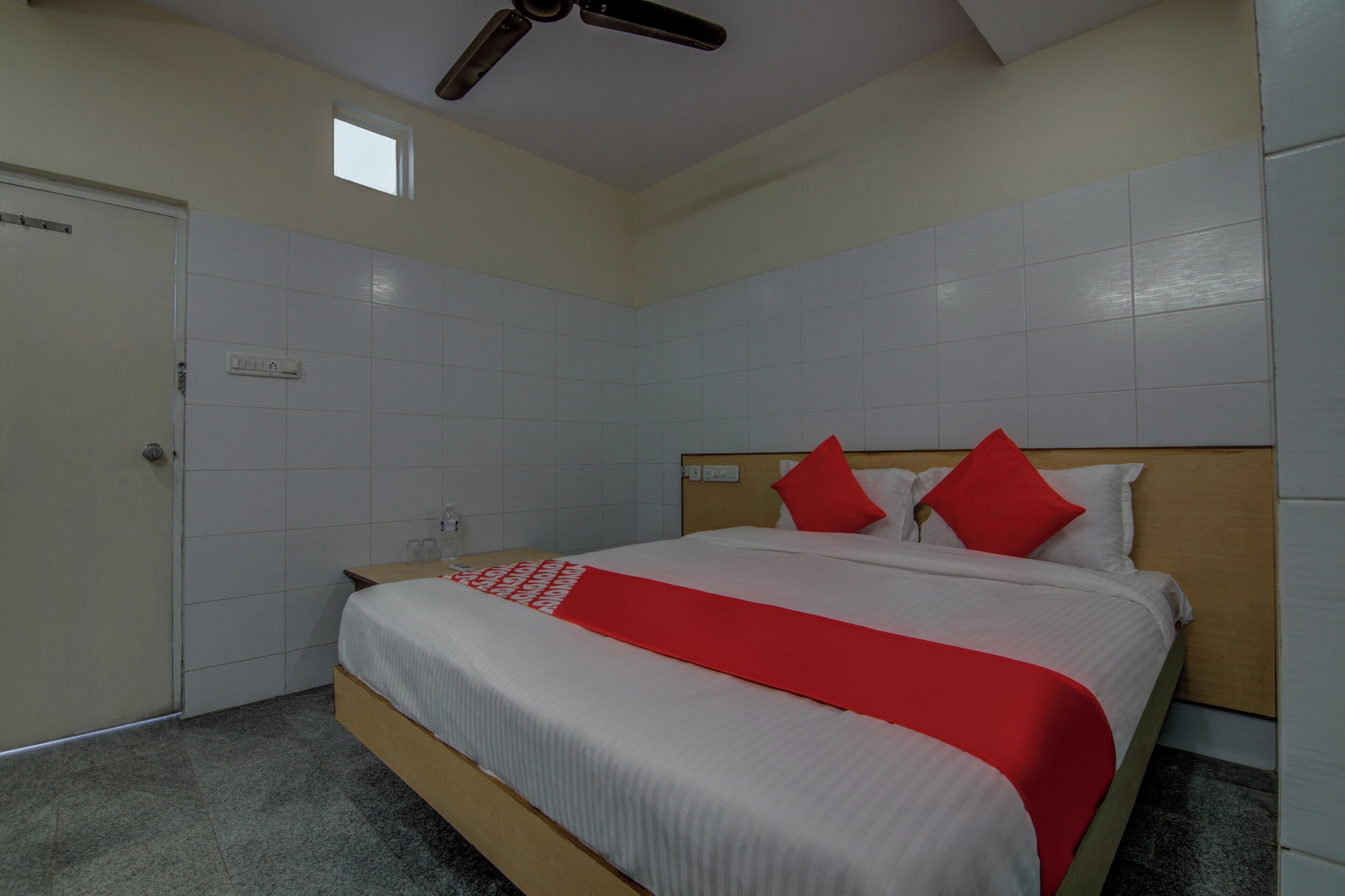 A hotel room with a bed, a ceiling fan, a small window, and a door.