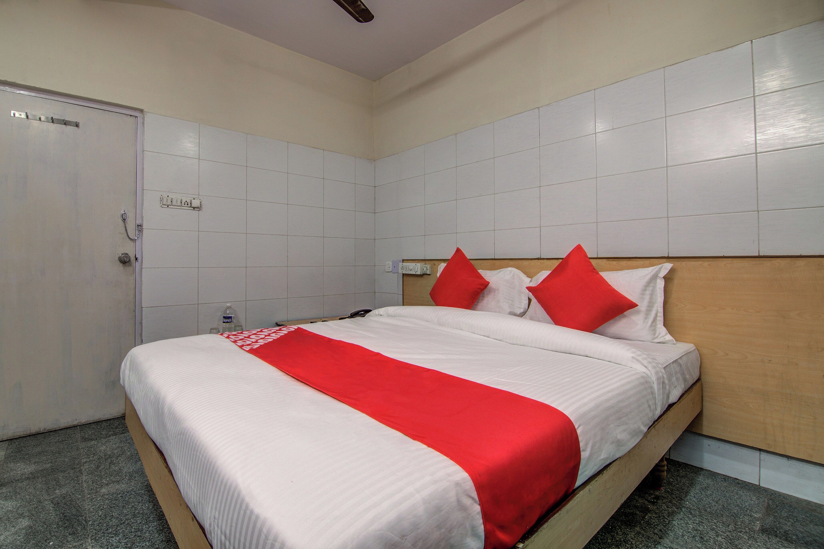 A hotel room with a bed, a wooden headboard, a white wall, a door, and a light switch.