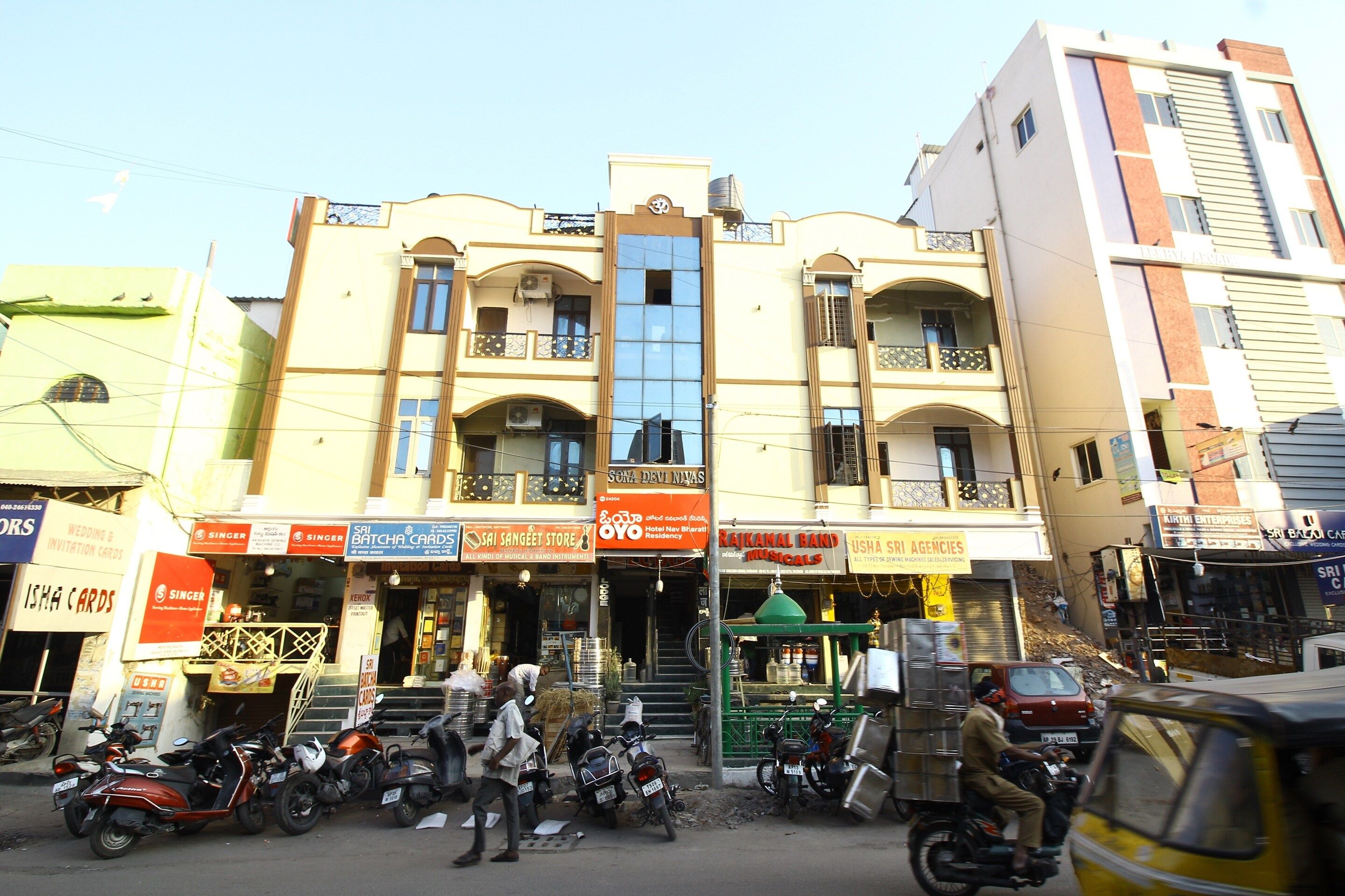 Un edifici de diverses plantes amb locals comercials a la planta baixa, incloent una botiga amb un cartell que diu 'SRI BATCHA CARDS' i una altra per a 'SRI SANGEET STORE'.