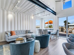 A modern living room with a sectional sofa, a dining table with chairs, and a large window with an umbrella. - Orange Sunset: Container Home w/ Heated Plunge Pool, Near Beach, Sleeps 10 (Port Aransas)