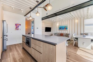 A modern kitchen with a central island, wooden cabinetry, and a dining area with a white table and chairs. - Orange Sunset: Container Home w/ Heated Plunge Pool, Near Beach, Sleeps 10 (Port Aransas)