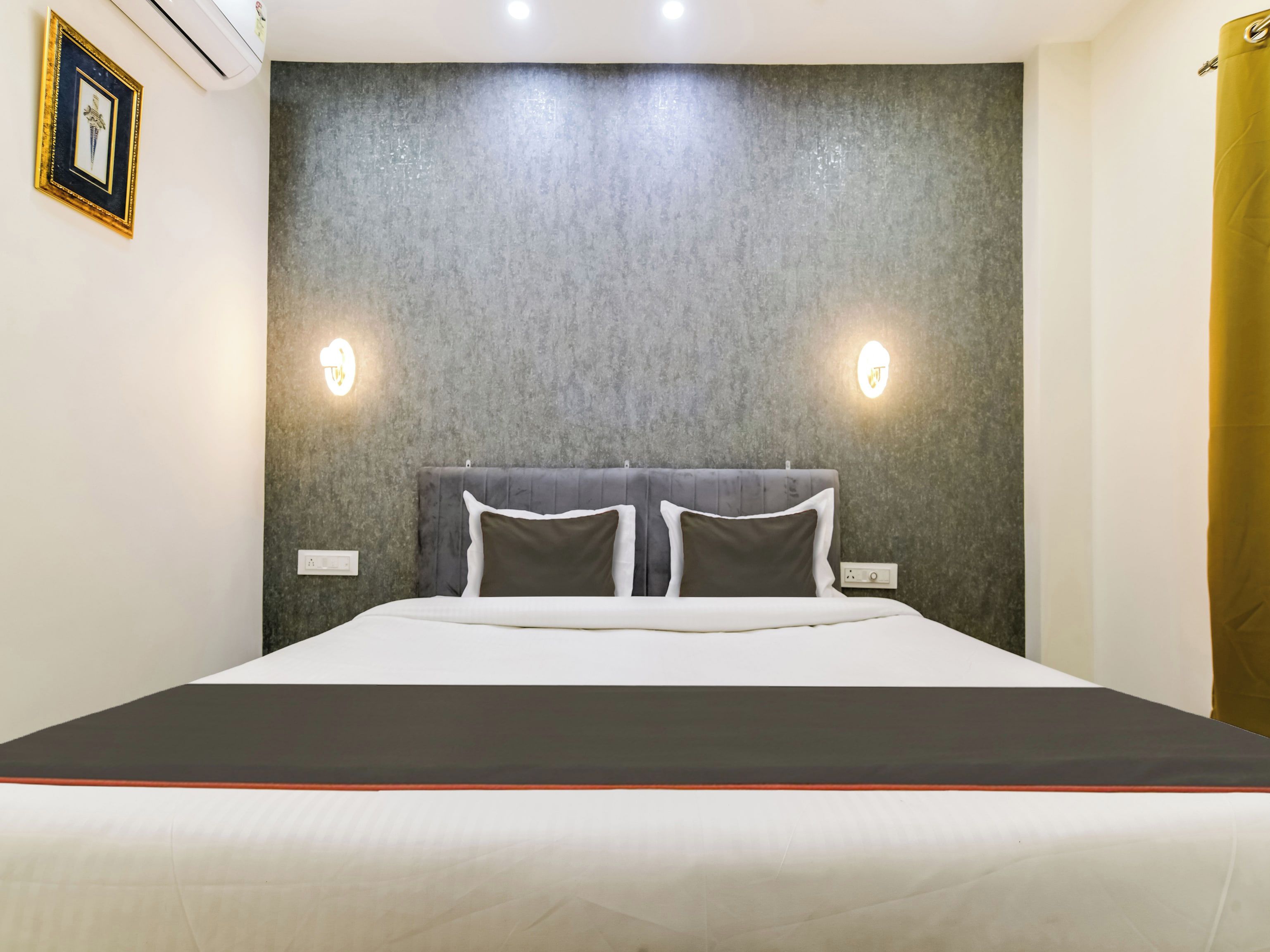 A hotel room with a large bed, grey textured wall, two wall-mounted lights, and a framed picture on the wall.