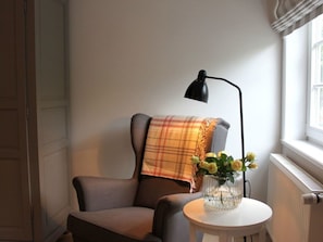 Una habitación con una silla, una mesa con un florero, una lámpara y un ventanal.