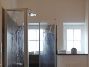 Eine begehbare Duschkabine mit Handbrause, ein Fenster mit Jalousien und ein Handtuchhalter mit gefalteten Handtüchern.