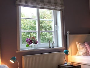 Ein Schlafzimmer mit Fenster, Heizkörper, Nachttisch mit Lampe und Bett mit Kissen.