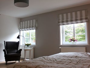 Un dormitorio con cama, una silla, una mesa con un florero y dos ventanas con cortinas.