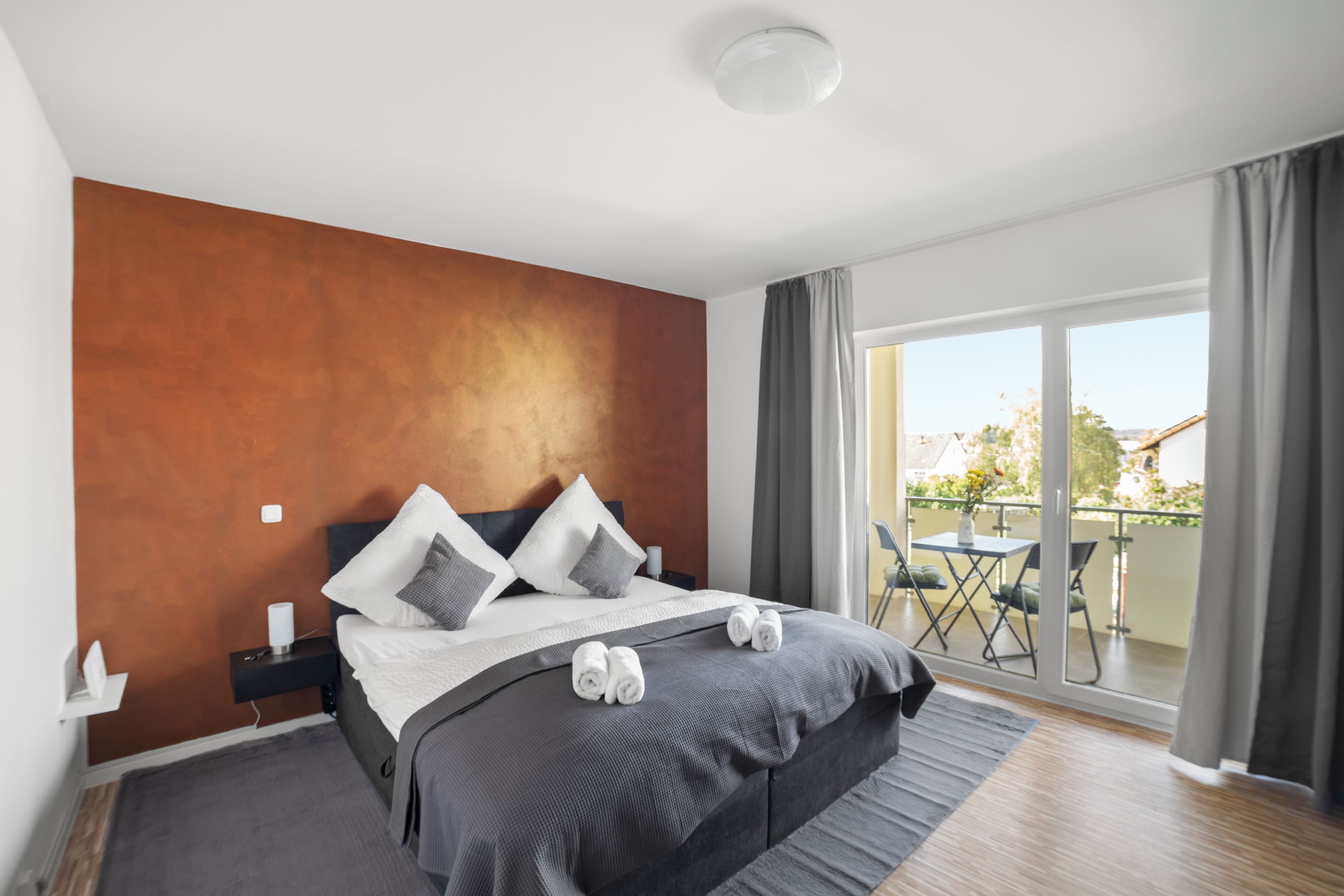 Une chambre moderne avec un grand lit, une table de chevet, une commode, une fenêtre avec des rideaux et un balcon agrémenté de mobilier d’extérieur.