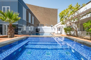 A swimming pool with blue mosaic tiles, surrounded by a modern building with white and grey walls, and a palm tree. - High-Floor 2BR with City Views and Near Dubai's Major Landmarks, Sky Gardens (Dubai)