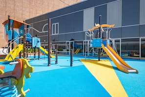 A playground with various play structures, including slides and climbing frames, in front of a building with large windows. - High-Floor 2BR with City Views and Near Dubai's Major Landmarks, Sky Gardens (Dubai)
