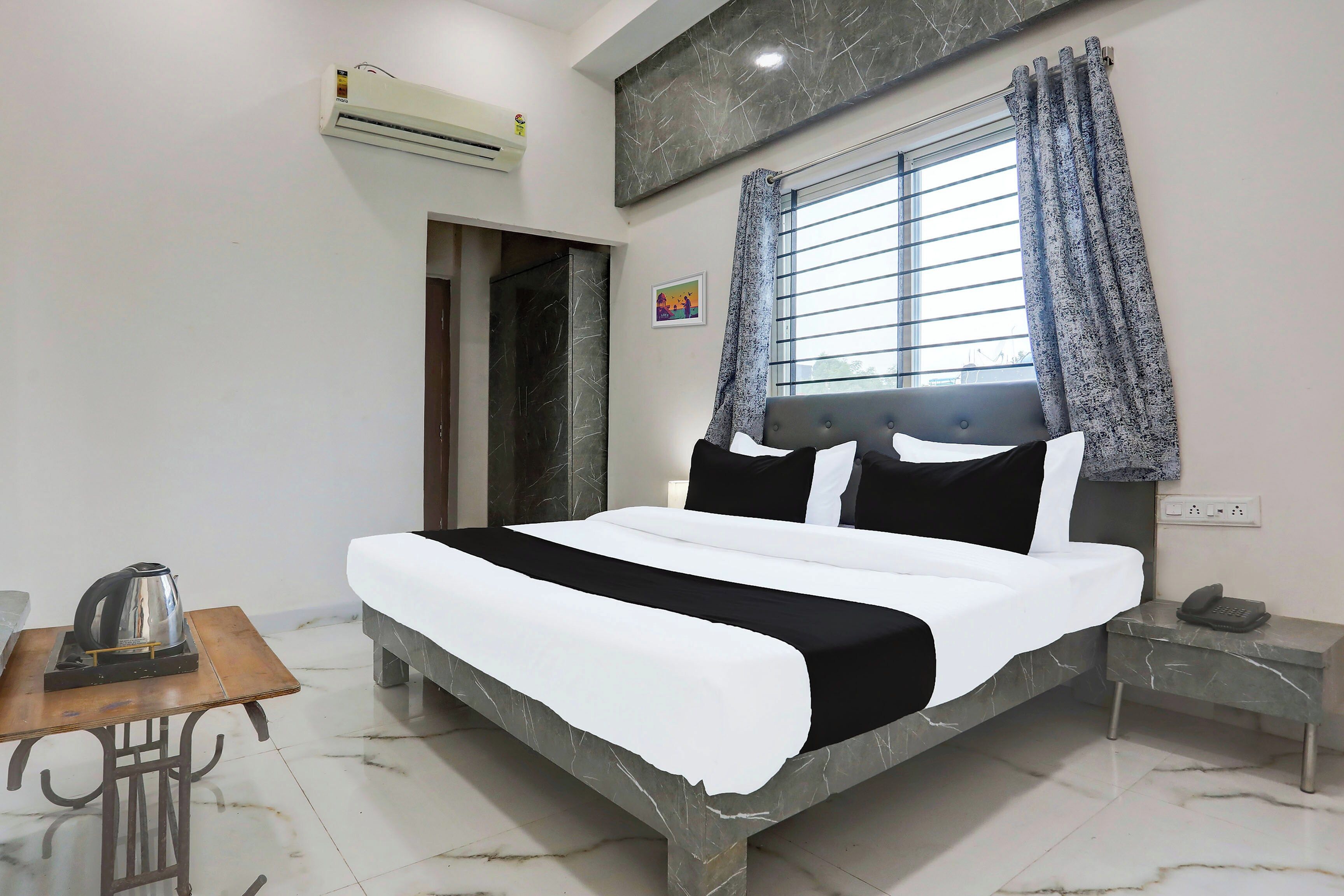 A hotel room with a bed, a bedside table with a phone, a kettle, and a picture on the wall.