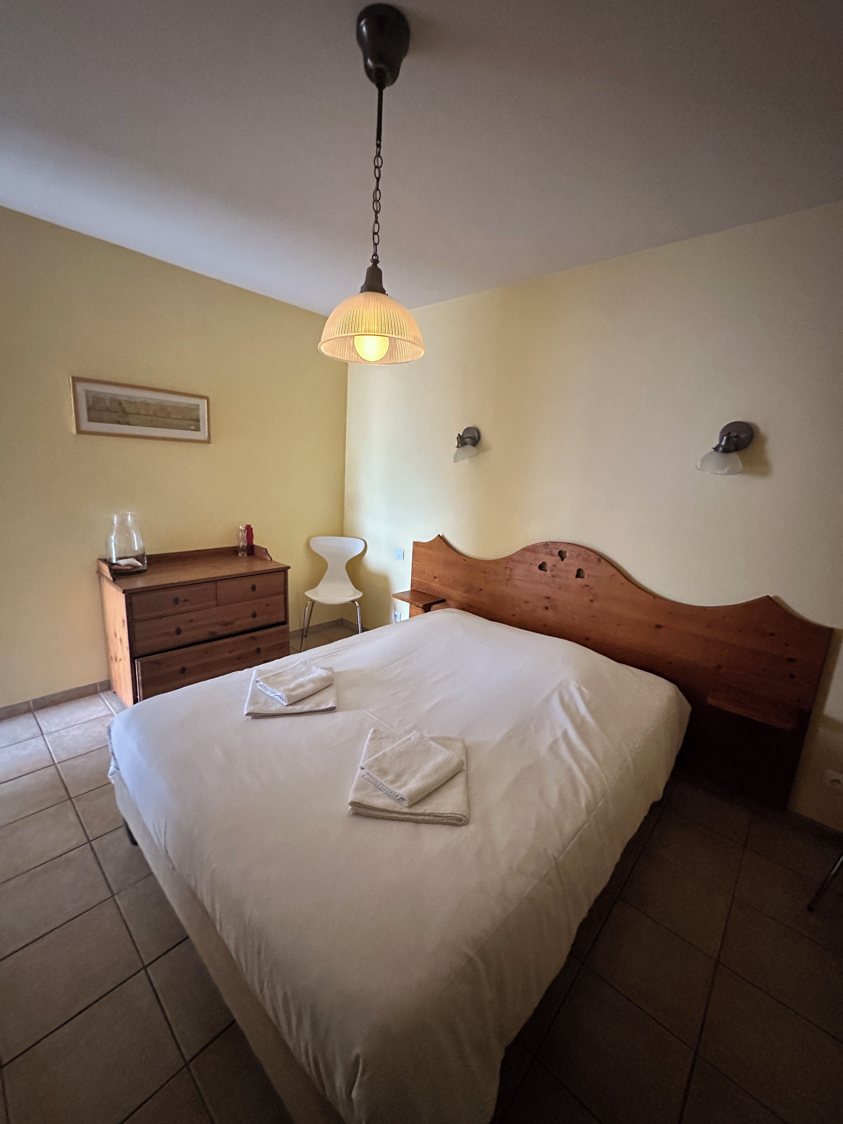 A hotel room with a bed, a wooden dresser, a chair, and a hanging light fixture.