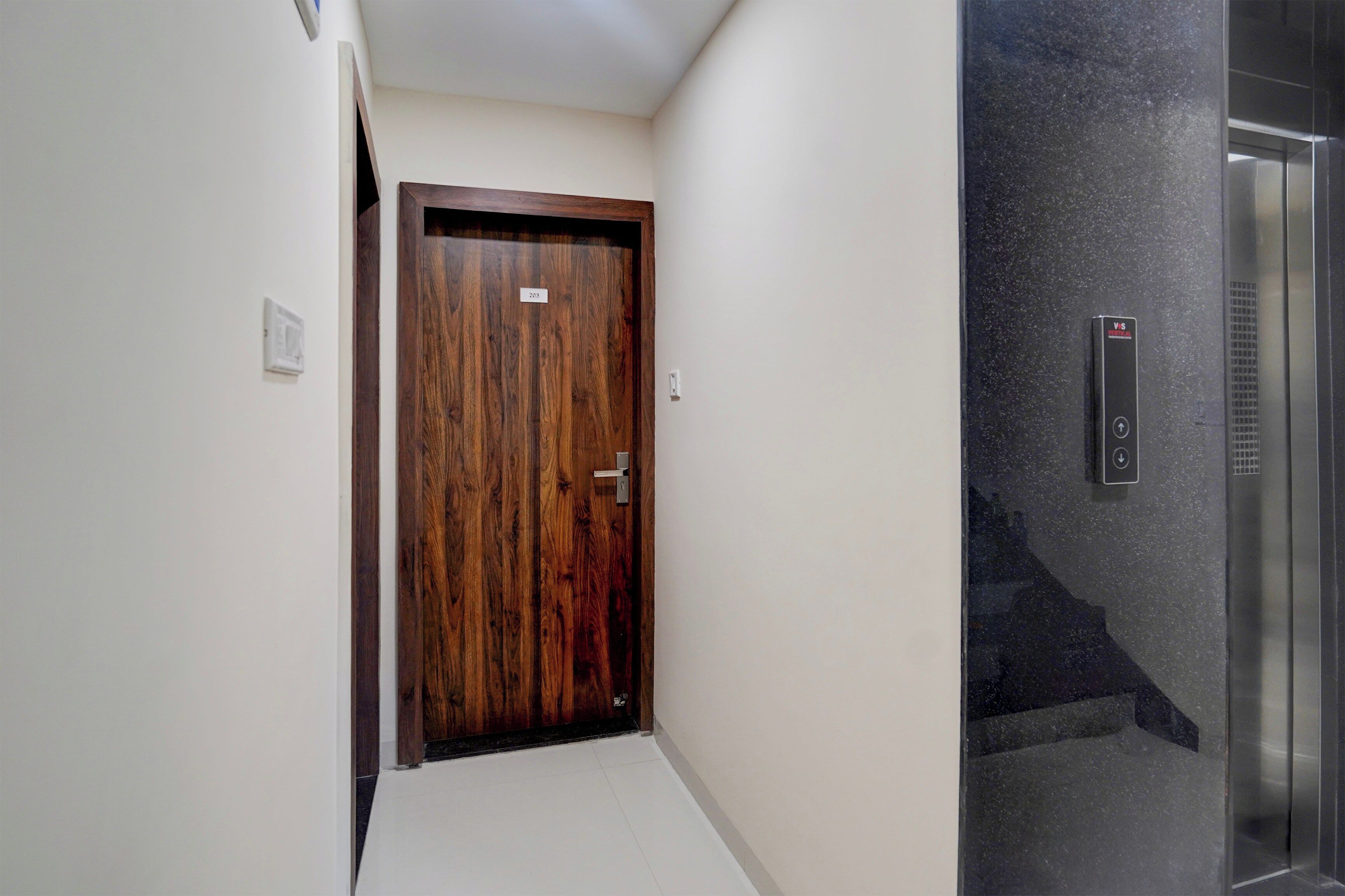 A hallway with a wooden door, a wall-mounted elevator control panel, and a tiled floor.