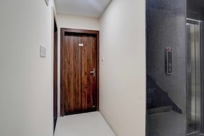 Un pasillo con una puerta de madera, un panel de control de ascensor en la pared y un suelo de baldosas.