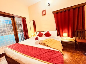 Een hotelkamer met een groot bed, rode gordijnen, een houten hoofdeinde, een nachtkastje met een lamp en uitzicht op een zwembad door een raam met metalen spijlen.