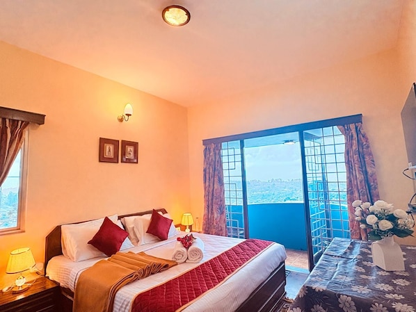 A hotel room with a bed, a nightstand, a table with a vase, and a view of the city through a large window. - Aura Solace Home Stay (Kodaikanal)