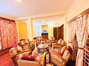 A spacious living room with a round coffee table surrounded by wicker chairs and sofas, a television, and a wooden cabinet. - Aura Solace Home Stay (Kodaikanal)