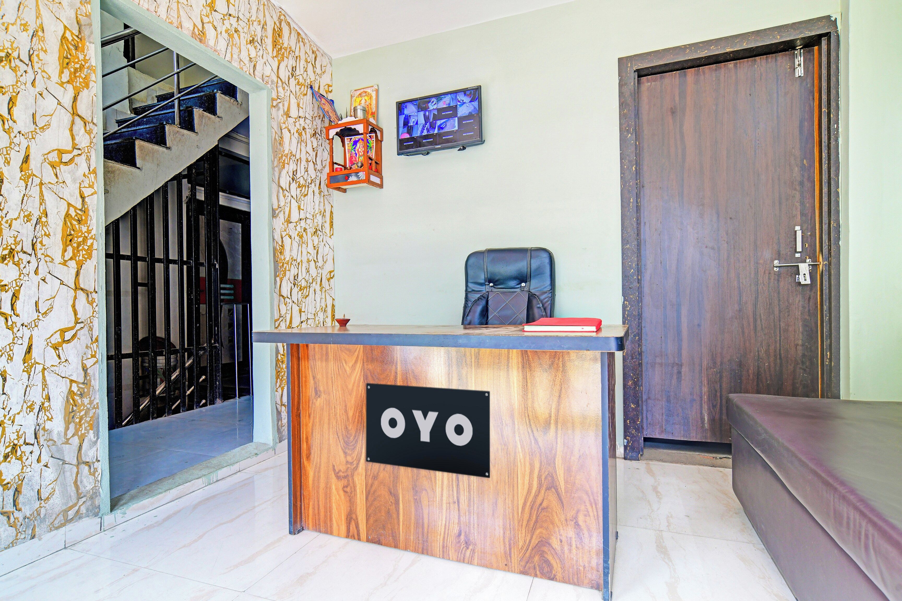 A reception desk with a sign reading 'OYO', a staircase, a door, and a wall-mounted television.
