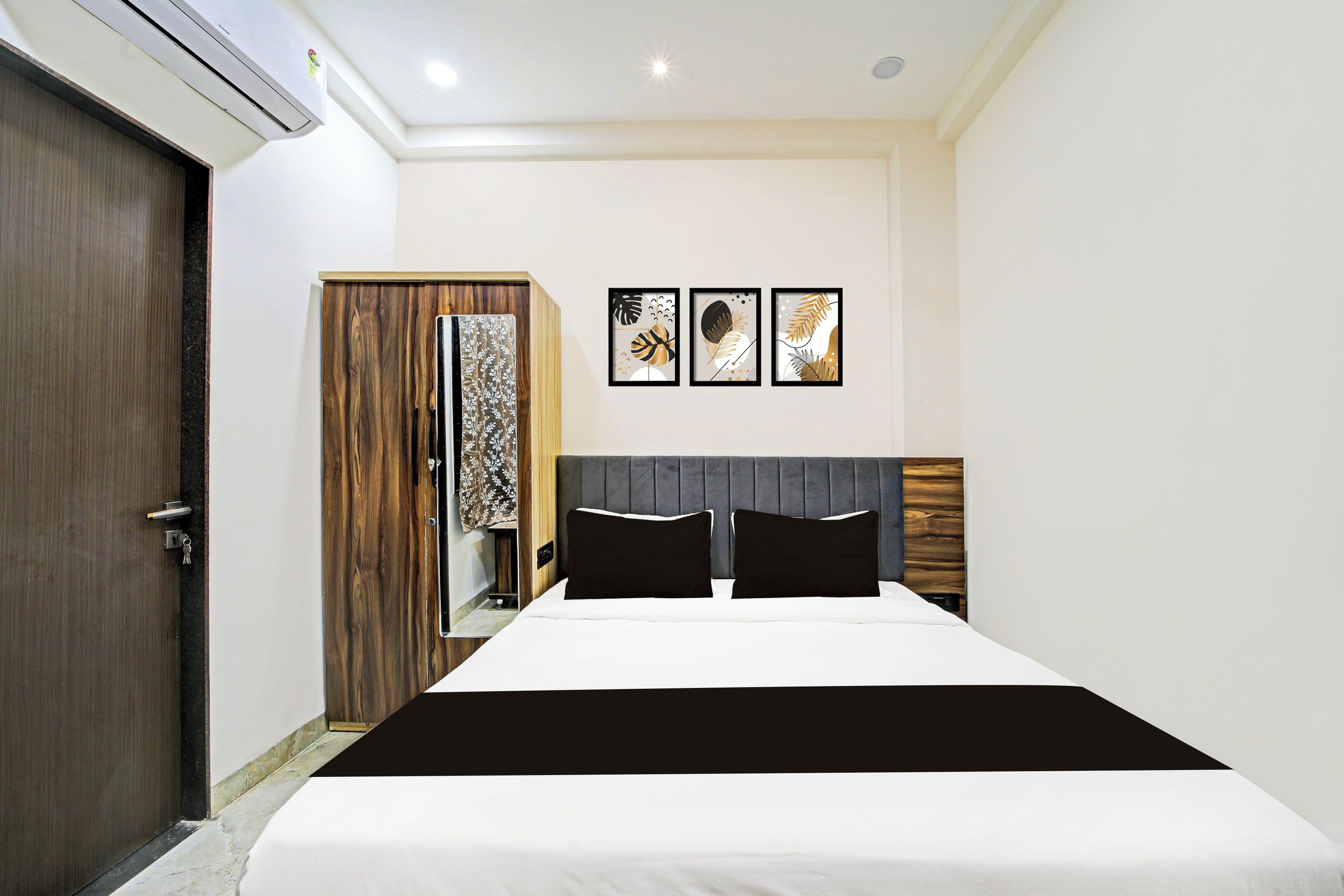 A hotel room with a bed, a wardrobe, a mirror, and three framed artworks on the wall.