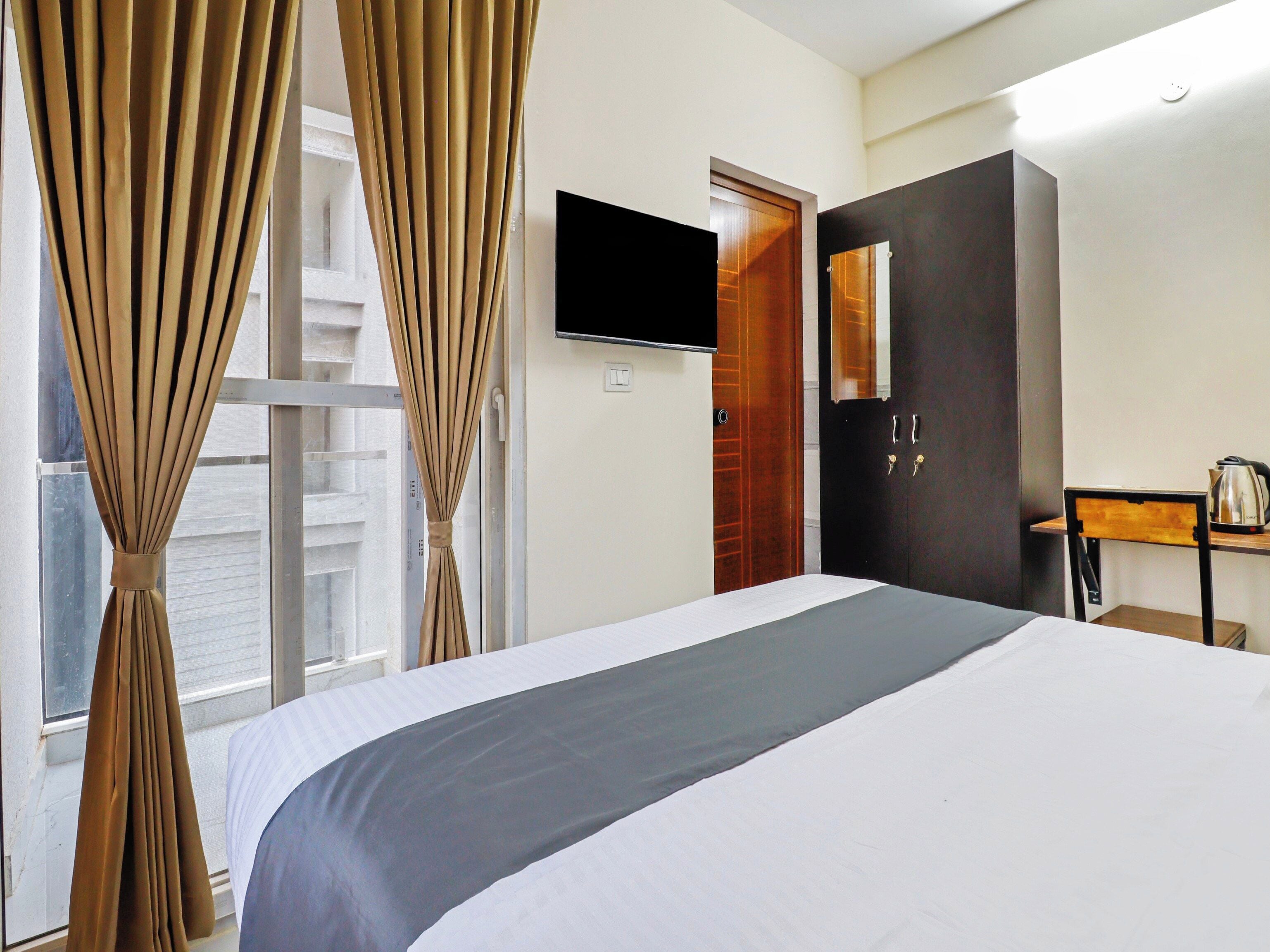 Habitación de hotel con cama, cortinas, televisor, armario y una mesita con hervidor.