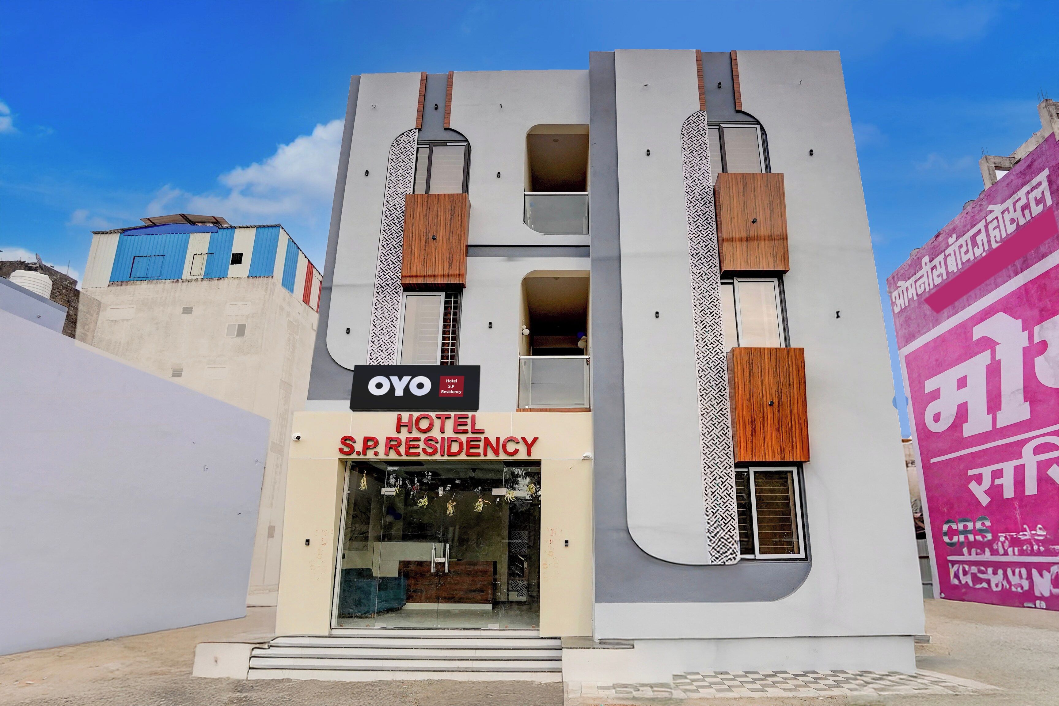 Um hotel moderno com uma placa grande de 'OYO Hotel S.P. Residency', uma entrada espaçosa e uma fachada marcante com ripas verticais de madeira.