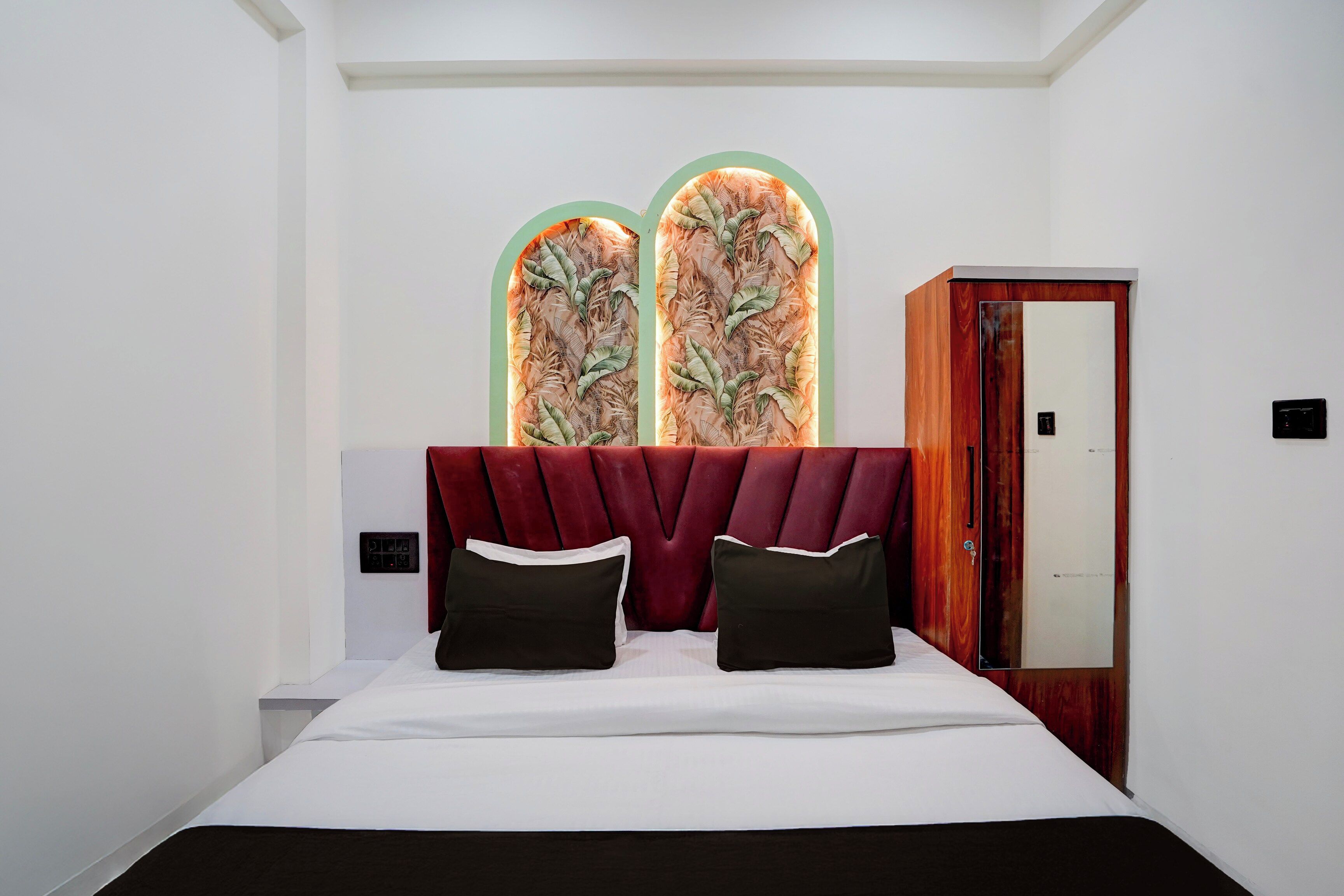 Un dormitorio con cama, dos almohadas, un cabecero tapizado en rojo, un armario de madera y un interruptor de luz en la pared.