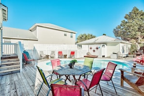 A pool area with a wooden deck, outdoor furniture, and a white building in the background. - A Wave From It All-crystal Beach (Destin)