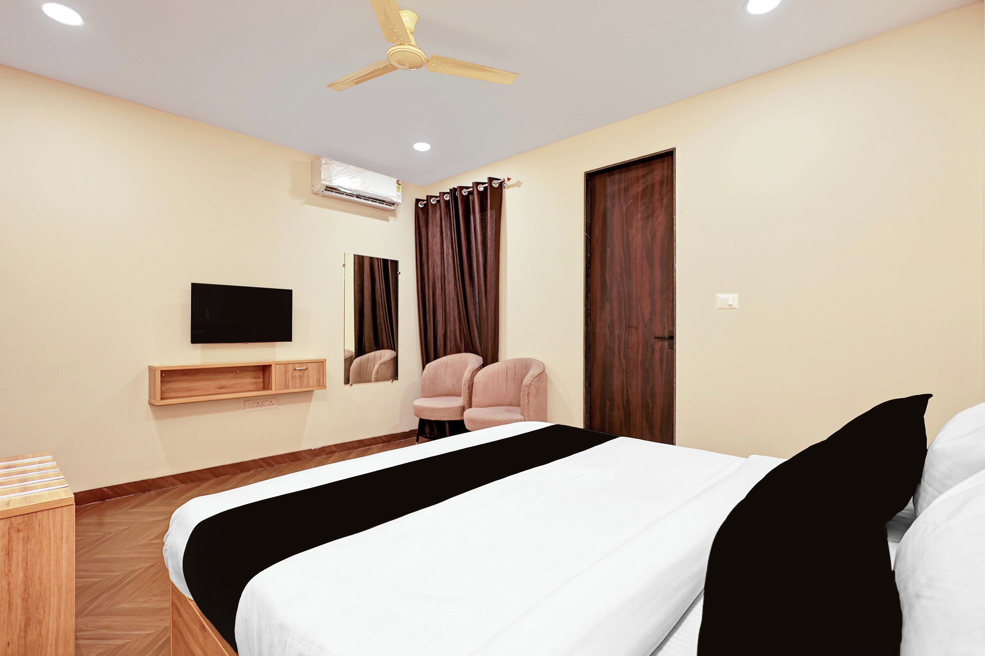 Quarto de hotel com cama, televisão, ventoinha no teto, ar condicionado, espelho e porta.