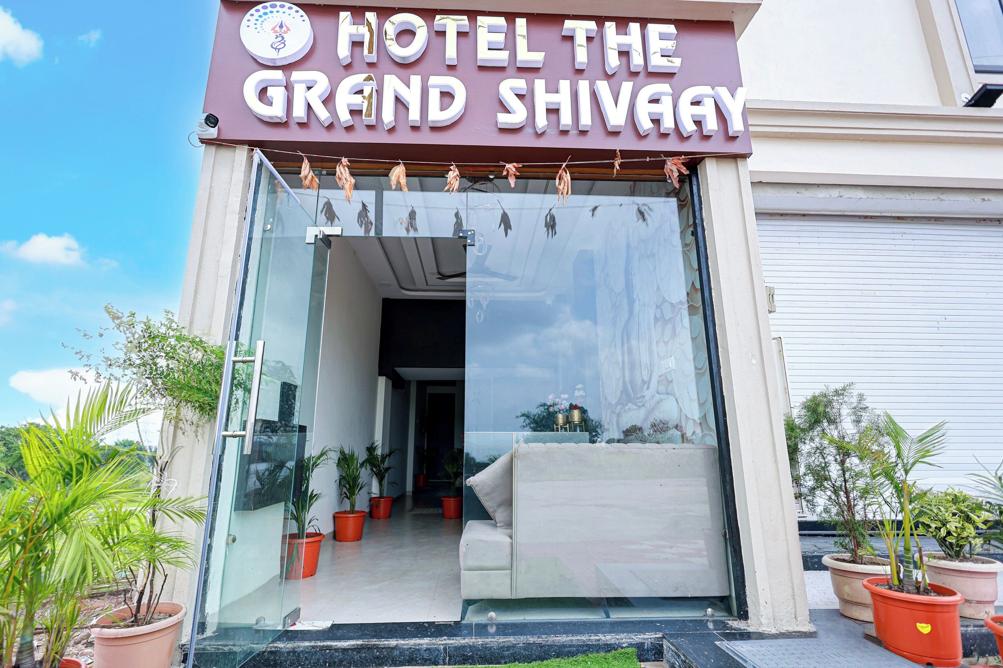 La entrada al Hotel The Grand Shivay, con fachada de vidrio, plantas en macetas y un interior visible.