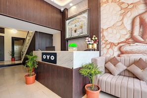 Una mesa de recepción con un cartel que dice 'OYO', flanqueada por plantas en macetas, un sofá con cojines y una pared decorativa con un motivo de hojas.