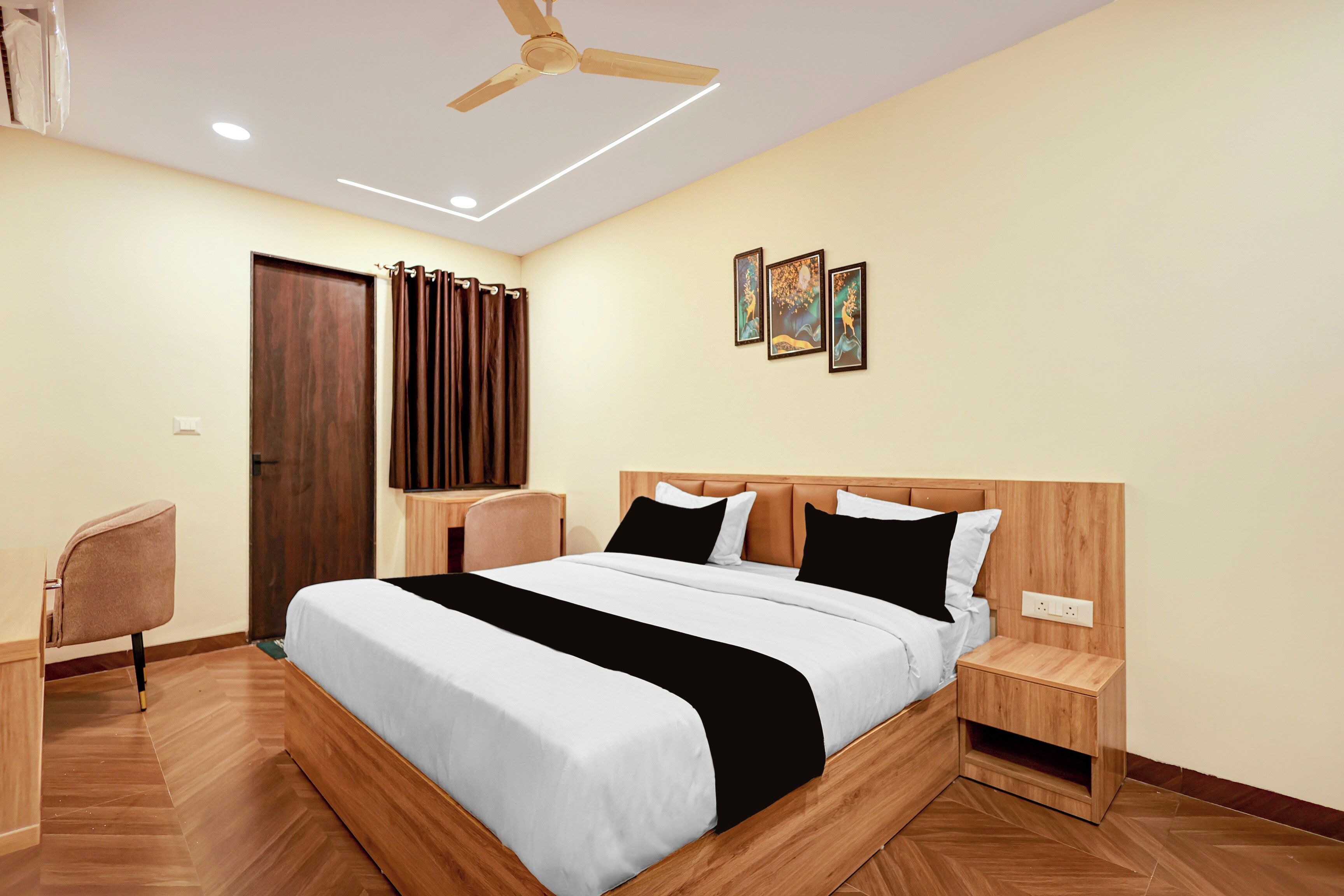 Habitación de hotel con cama, mesitas de noche, escritorio, silla, ventilador de techo y una obra de arte en la pared.