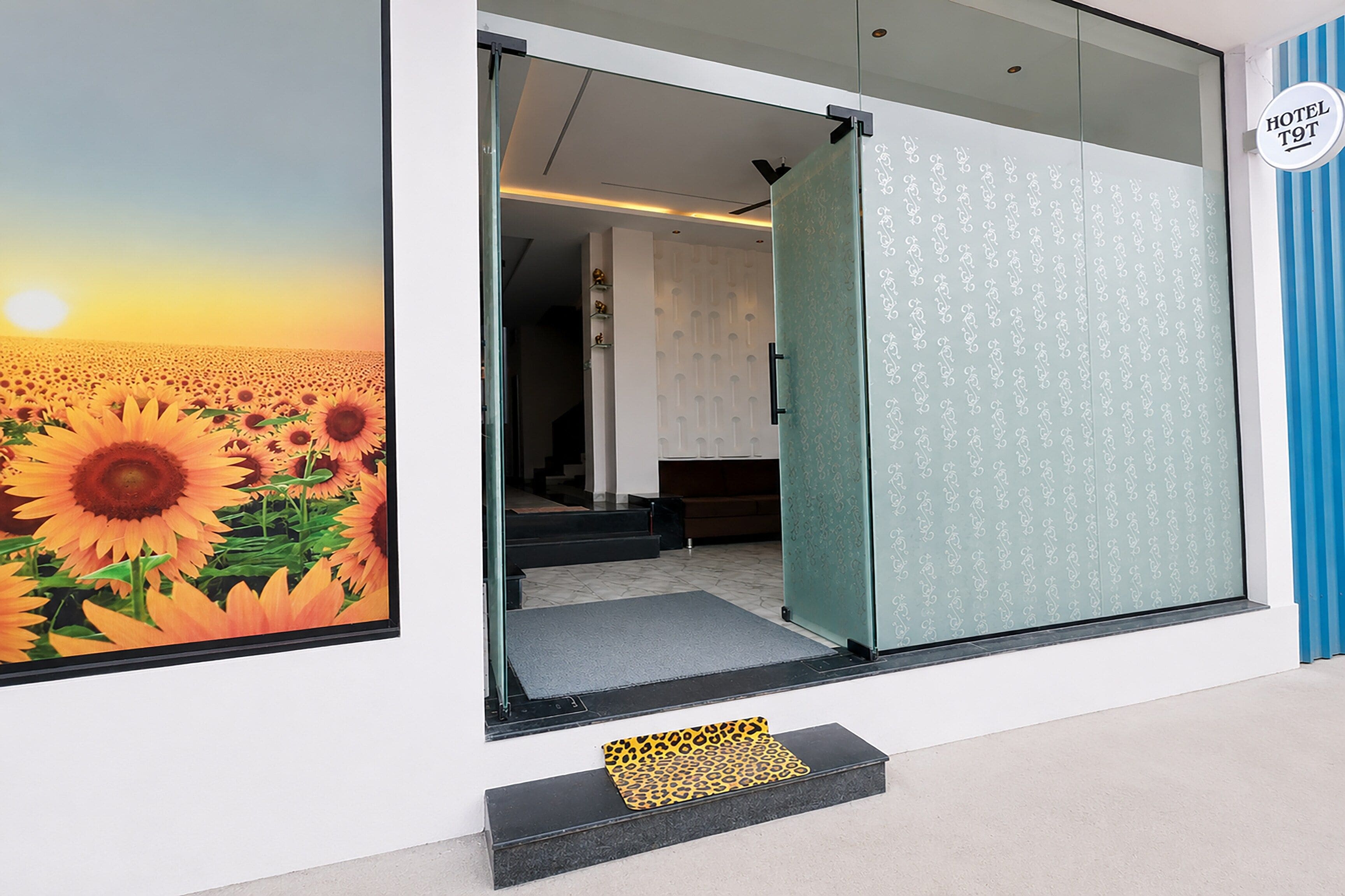 Entrada de un hotel con puerta de cristal, un mural de girasoles y una pared a rayas azules.