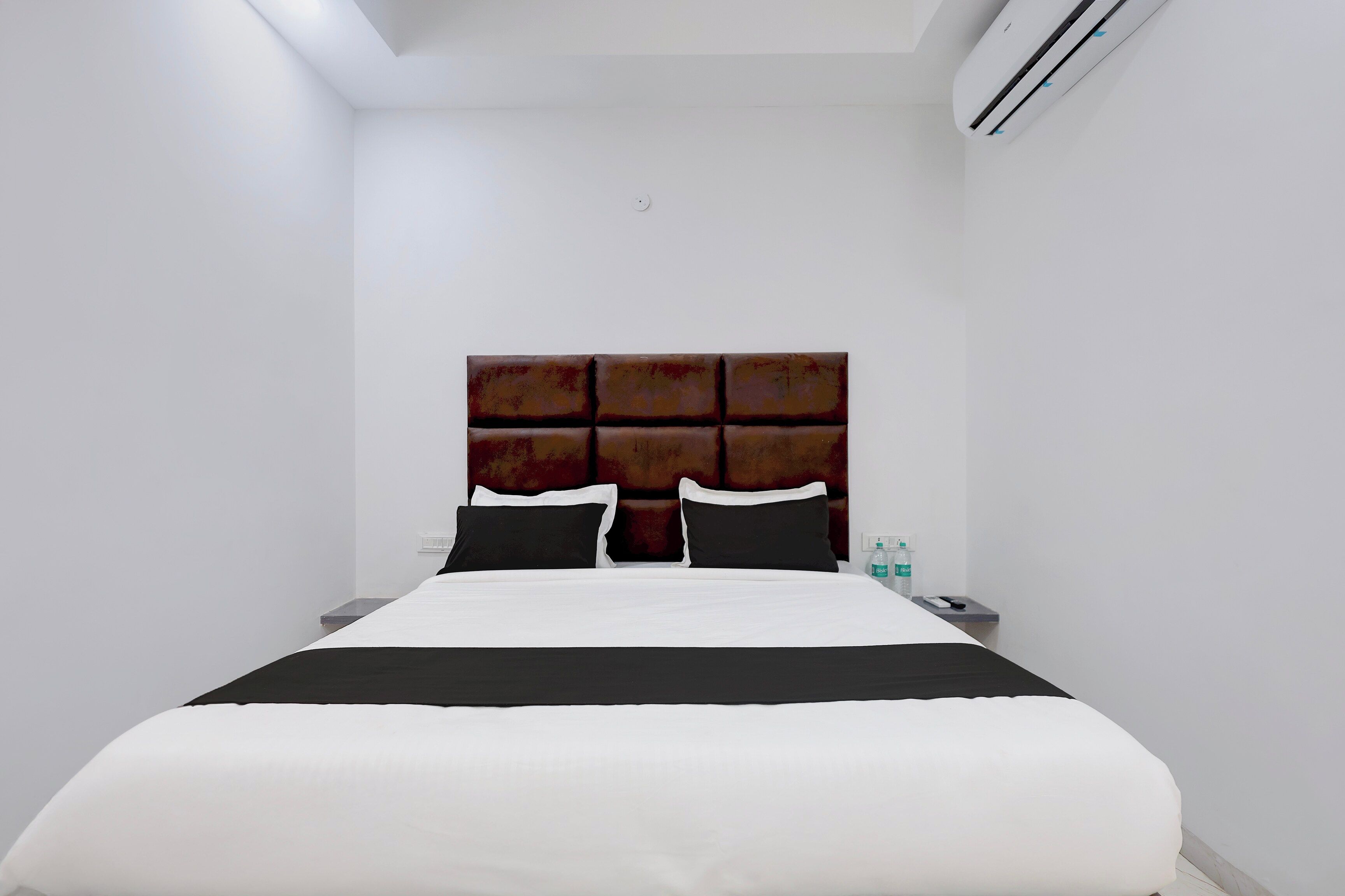 Una habitación de hotel con una cama, una mesita de noche, un equipo de aire acondicionado y una botella de agua.