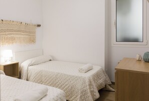 A bedroom with two beds, a bedside table with a lamp, a macrame wall hanging, and a window with a view.