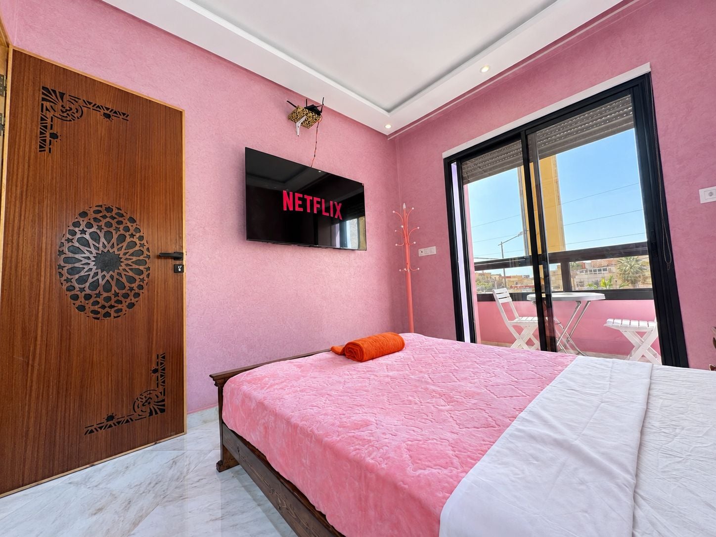 Een slaapkamer met een roze muur, een houten deur met een geometrisch ontwerp, een bed met een roze en witte dekbedovertrek, een televisie boven het bed waarop 'NETFLIX' staat, en een balkon met een tafel en stoelen.