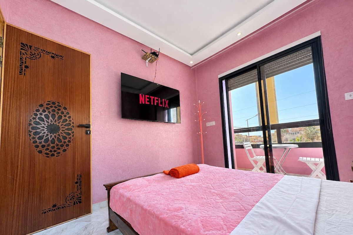 Un dormitori amb una paret rosa, una porta de fusta amb un disseny geomètric, un llit amb col·cha rosa i blanca, un televisor fixat sobre el llit que mostra 'NETFLIX', i un balcó amb una taula i cadires.