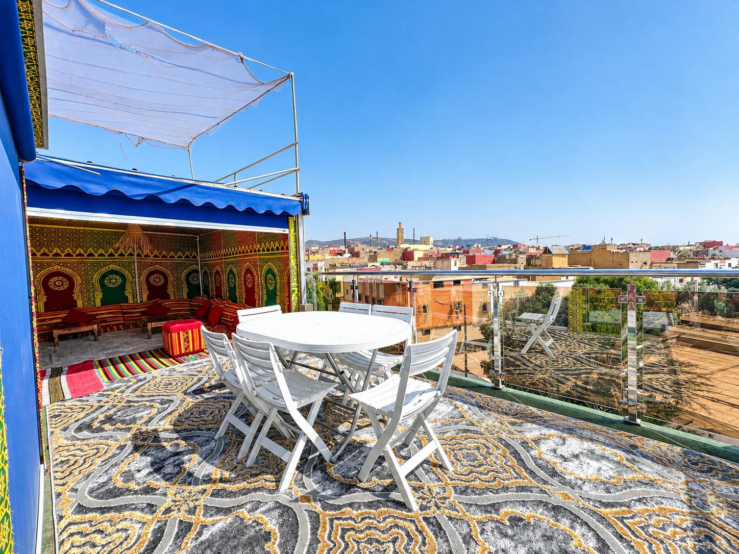 A rooftop terrace with a patterned floor, a table with chairs, and a seating area with cushions.