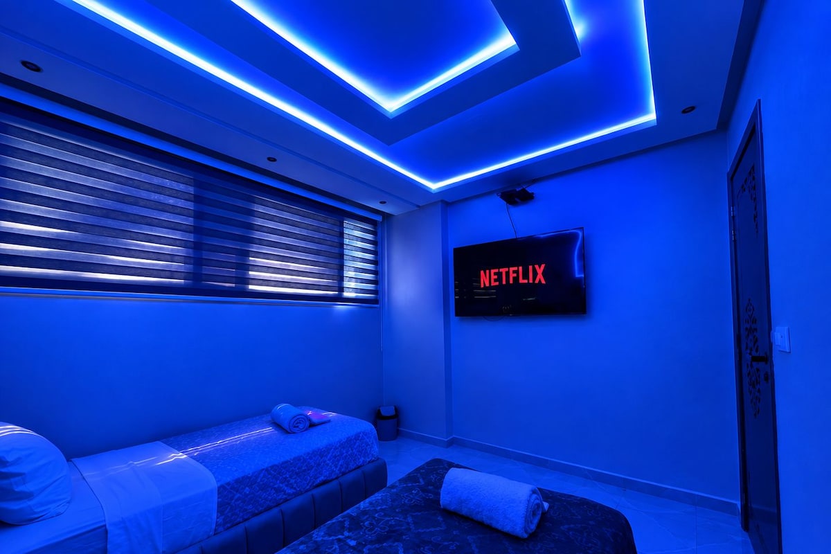 Una habitació amb llit, sofà, televisió penjada a la paret i finestra amb persianes.