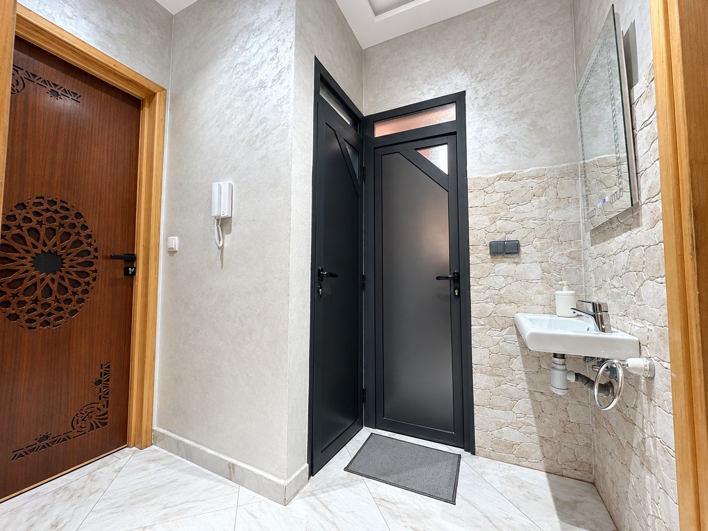 A bathroom with a black door, a wooden door with a decorative panel, a white sink with a stone backsplash, and a mirror above the sink.