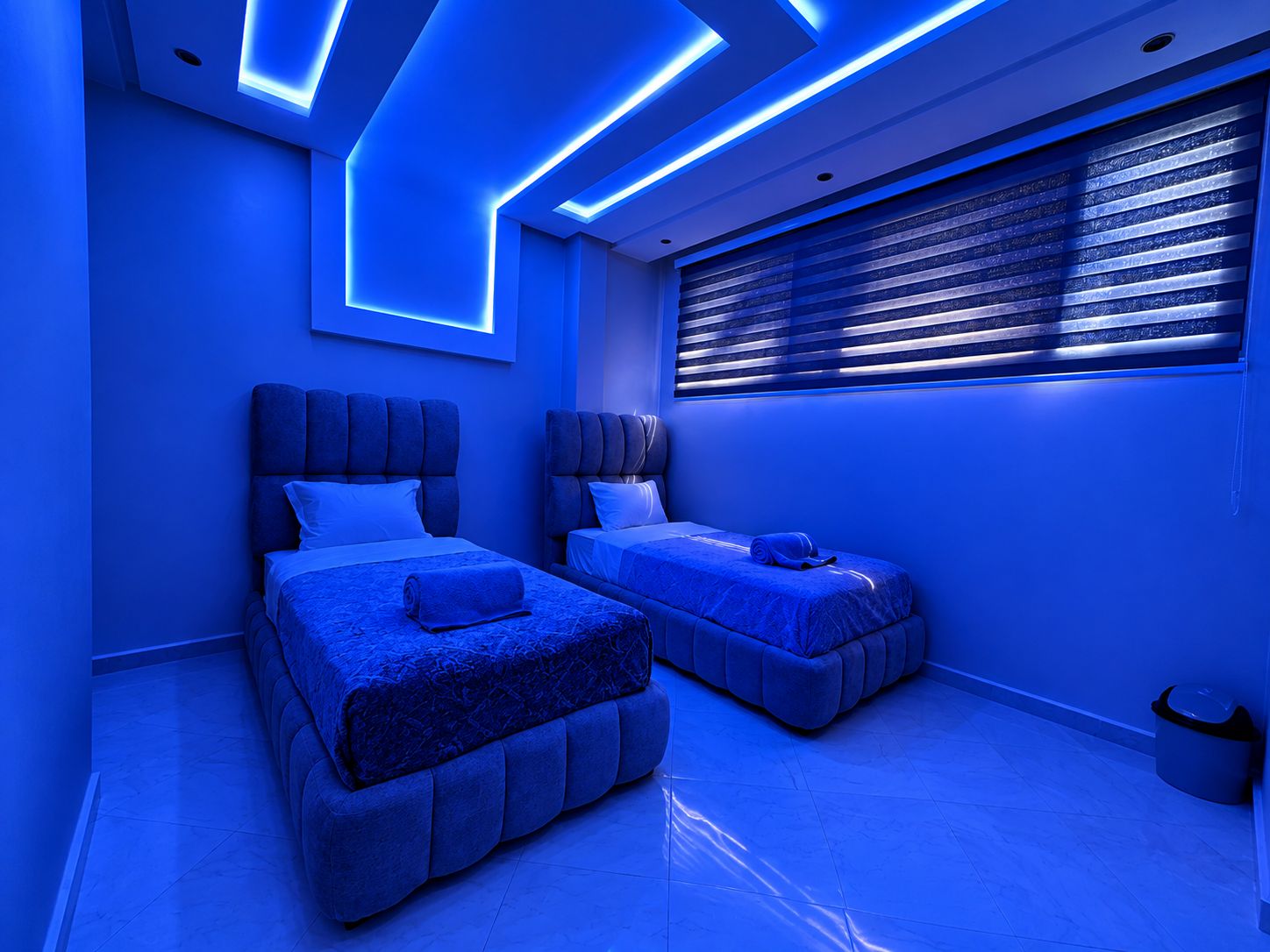 A modern bedroom with two beds, a large window with blinds, and a ceiling with blue lighting.