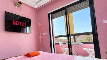A room with a bed, a TV mounted on the wall, and a balcony with chairs.